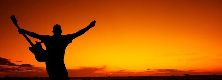 Musiker steht mit Gitarre vorm Sonnenuntergang - Musiklabel vs. Selbstvermarktung