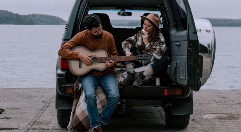 Eine Frau und ein Mann sitzen mit einer Decke im Kofferraum ihre Autos. Er spielt Gitarre, sie Mundharmonika