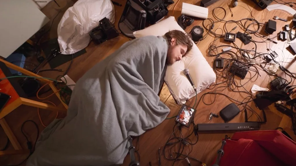 Bo Burnham liegt mit Bettwäsche auf dem Boden, um ihn herum liegt Technik, auf dem Kopfkissen ein Mikrofon.