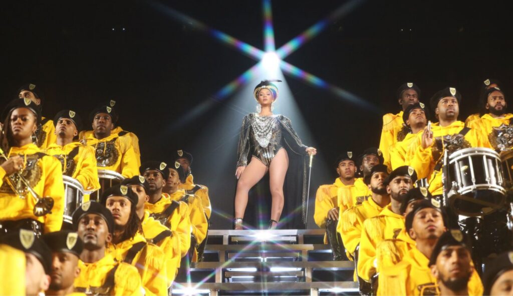 Beyoncé steht auf einer Bühnentreppe in einem glitzernden Einteiler und einem Gehstock, um sie herum eine Kapelle aus People of Colour