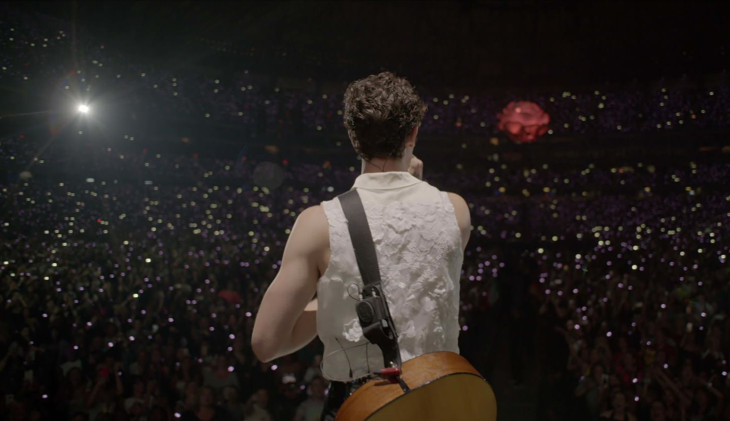 Shawn Mendes steht auf einer Bühne. Man sieht ihn von hinten mit seiner Gitarre umgeschnallt, das Publikum hält Lichter hoch