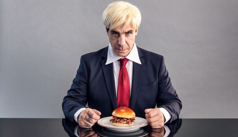 Rammstein-Sänger Till Lindemann sitzt mit blonder Perücke und ernstem Blick vor einem Teller mit einem Burger