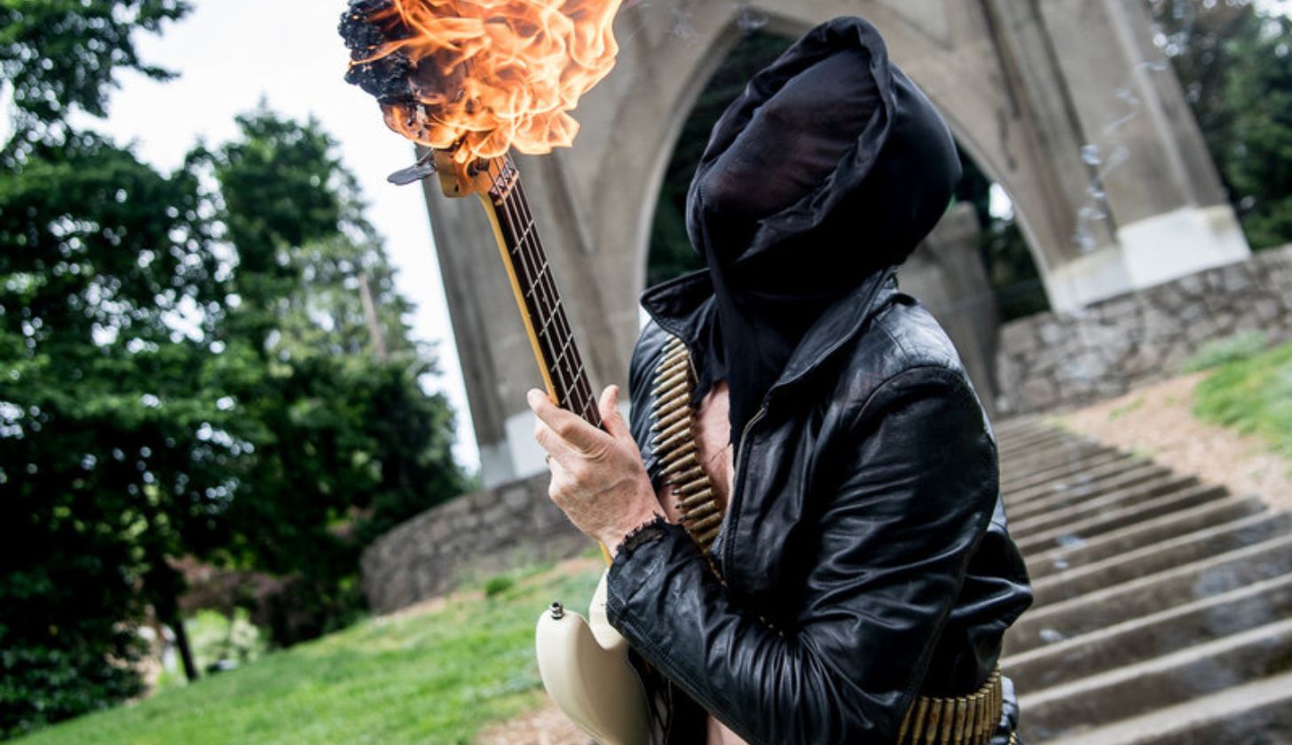 Ein Bandmitglied der Band Midnight in Lederjacke, schwarzer Maske und Patronengürtel hält einen brennenden Bass