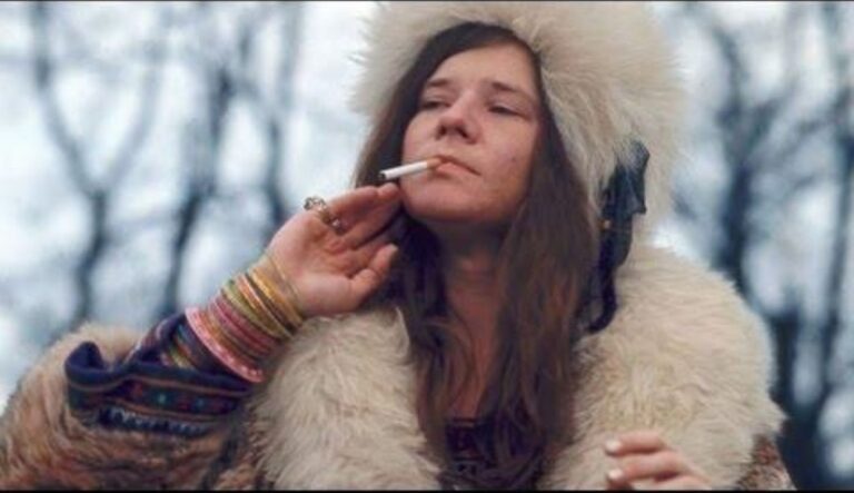 Ein Bild von Janis Joplin in dicker Jacke und Mütze wie sie eine Zigarette raucht